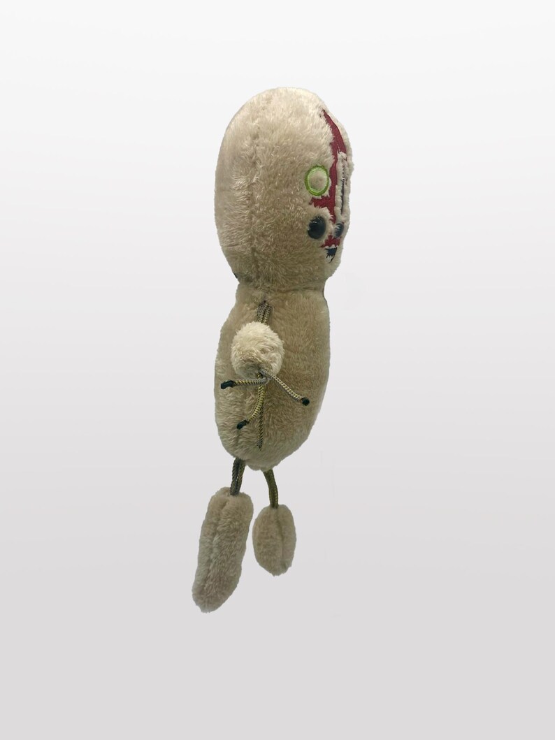 Puede incluir: Un juguete de peluche de una figura humanoide con una cara roja y blanca, ojos verdes y pelaje marr&oacute;n. El juguete tiene cuatro extremidades y est&aacute; de pie sobre un fondo blanco.