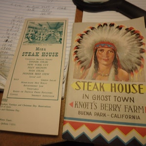 May include: A vintage menu for the Steak House in Ghost Town at Knott's Berry Farm in Buena Park, California. The menu features a charcoal broiled steak dinner with a variety of sides, including green salad, home-made hot rolls, and French fried potatoes.