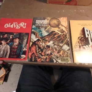 May include: Three vintage movie promotional paperbacks. The first is red with the title "OLIVER!" and a photo of two boys. The second is a colorful image of a volcano erupting with the title "Krakatoa East of Java in Cinerama". The third is yellow with the title "The Story of the Making of Ben-Hur" and a photo of a chariot race.