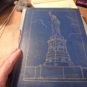 May include: A blue hardcover book with a gold embossed image of the Statue of Liberty on the front cover.