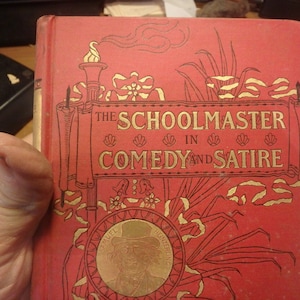 May include: The cover of a red book titled "The Schoolmaster in Comedy and Satire" with gold lettering and illustrations. The book features a portrait of Samuel Butler.