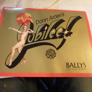 May include: A vintage poster with a gold background and a red border. The poster features the text "Donn Arden's Jubilee" in black script, a showgirl illustration, and the Bally's Las Vegas Nevada logo.