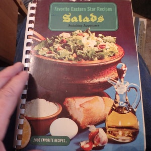 May include: A vintage cookbook with a spiral binding titled "Favorite Eastern Star Recipes Salads Including Appetizers". The cover features a colorful photo of a salad in a wooden bowl, a loaf of bread, a garlic clove, an egg, and a glass bottle of oil.