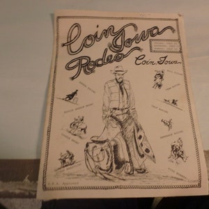 May include: A black and white illustration of a cowboy riding a horse. The illustration is surrounded by a rope border and includes the text "Coin Iowa Rodeo Coin, Iowa".