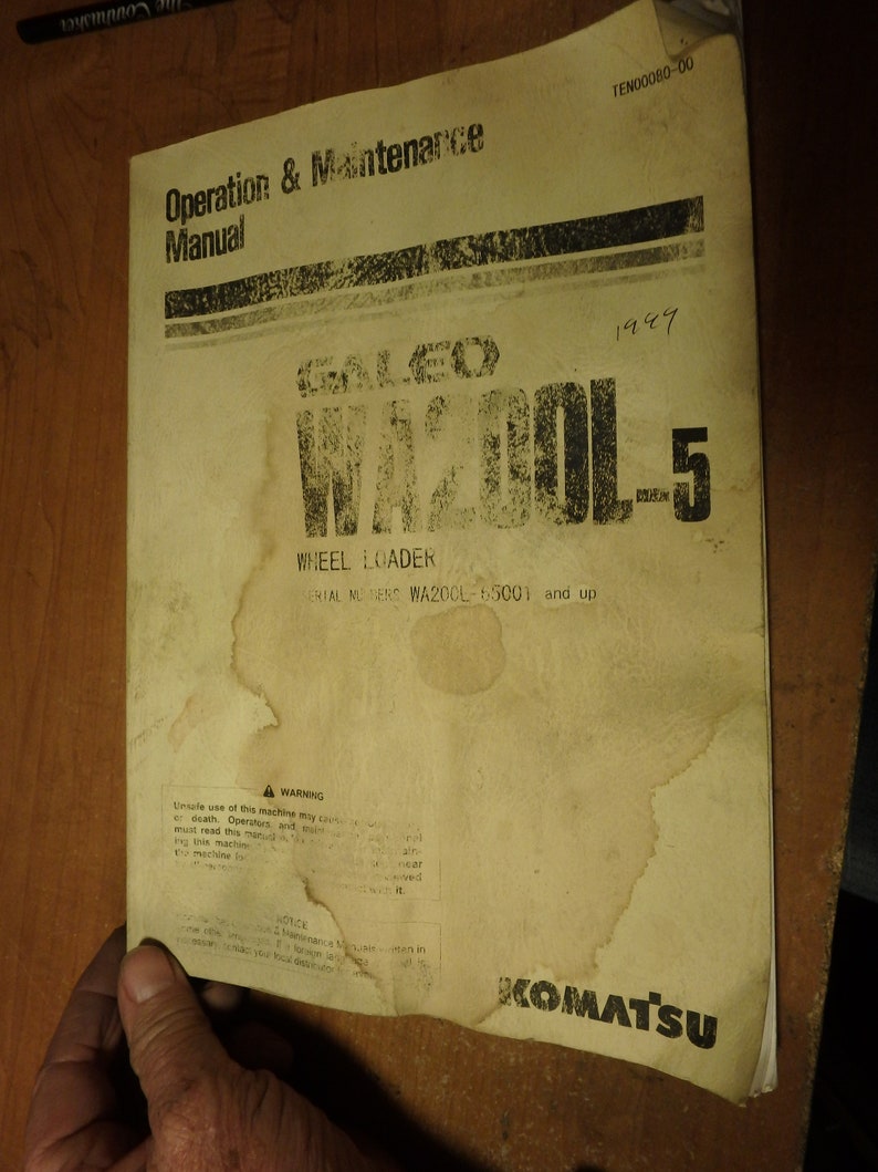May include: A yellowed, worn manual for a Komatsu WA200L-5 wheel loader. The manual has a warning sign and text that reads "Operation & Maintenance Manual" and "Serial Numbers WA200L-65001 and up".