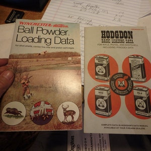 May include: Two vintage reloading data manuals. The left manual is a Winchester-Western Ball Powder Loading Data manual with a hunting scene on the cover. The right manual is a Hodgdon Basic Loading Data manual with four cans of Hodgdon powder on the cover.