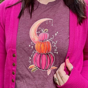 May include: A maroon t-shirt with a graphic of three stacked pumpkins with a crescent moon behind them. The pumpkins are pink, orange, and a darker pink. The graphic is surrounded by vines and stars.
