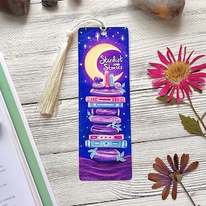 May include: A purple and blue bookmark with a crescent moon and stars. A stack of books is on top of a pile of stones. The text "Stardust and Stories" is on the bookmark.