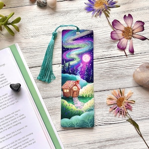 May include: A rectangular bookmark featuring a vibrant illustration of a small cottage with a glowing window, a stone path, and a bright full moon set against a dark night sky with a swirling purple and green aurora borealis and a teal tassel.