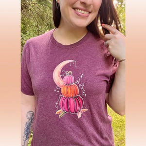 Puede incluir: Una camiseta color granate con un gr&aacute;fico de tres calabazas apiladas en tonos rosa, naranja y melocot&oacute;n. Las calabazas est&aacute;n sentadas sobre una luna creciente con enredaderas y estrellas.