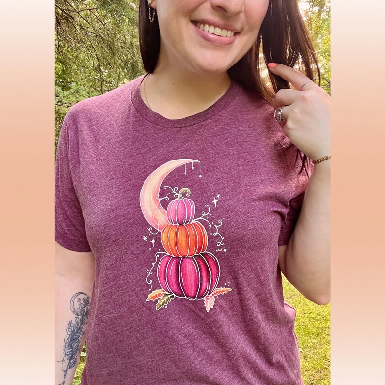 Puede incluir: Una camiseta color granate con un gr&aacute;fico de tres calabazas apiladas en tonos rosa, naranja y melocot&oacute;n. Las calabazas est&aacute;n sentadas sobre una luna creciente con enredaderas y estrellas.