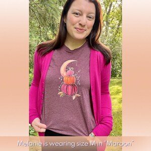 Puede incluir: Una camiseta color granate con un gr&aacute;fico de una pila de calabazas sobre una luna creciente. Las calabazas son naranja, melocot&oacute;n y rosa. La luna es blanca con un contorno dorado. El gr&aacute;fico est&aacute; rodeado de peque&ntilde;as estrellas blancas. "Melanie lleva la talla M en "Maroon"" est&aacute; escrito debajo de la imagen.