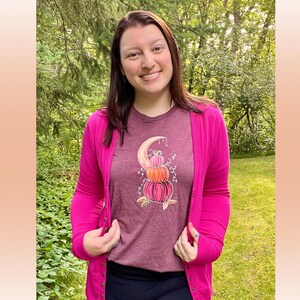 Puede incluir: Una camiseta granate con un gr&aacute;fico de una pila de calabazas con una luna creciente y estrellas. La camiseta la lleva puesta una persona con un c&aacute;rdigan rosa.