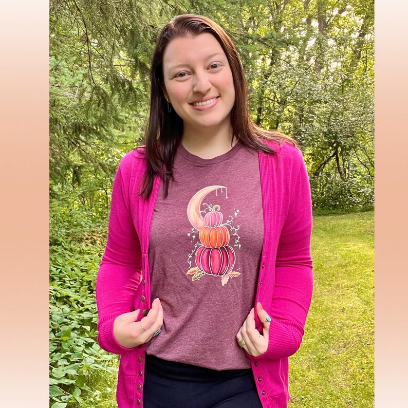 Puede incluir: Una camiseta granate con un gr&aacute;fico de una pila de calabazas con una luna creciente y estrellas. La camiseta la lleva puesta una persona con un c&aacute;rdigan rosa.