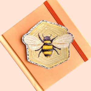 May include: A decorative bee-themed sticker with a silver glitter border, set on a light orange notebook. The bee has yellow and black stripes, with detailed wings. The notebook has an orange elastic band.