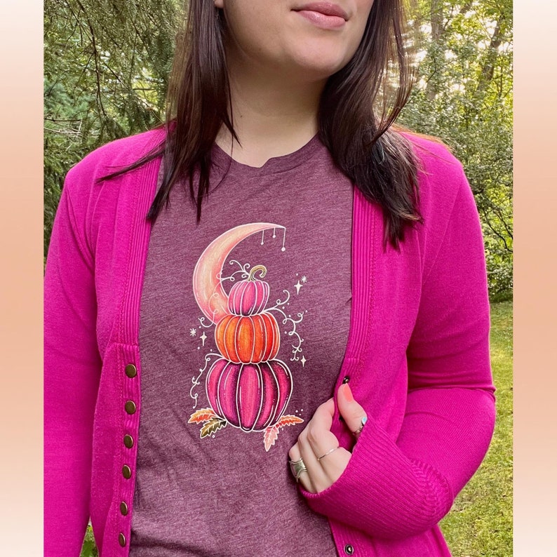May include: A maroon t-shirt with a graphic of three stacked pumpkins with a crescent moon behind them. The pumpkins are pink, orange, and a darker pink. The graphic is surrounded by vines and stars.
