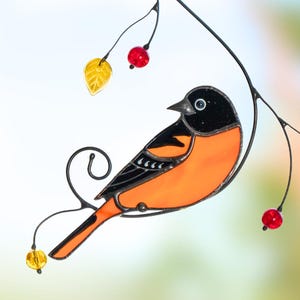 Brightly lit stained glass oriole with red beads and yellow leaves, hanging from a suction cup against a soft natural background.