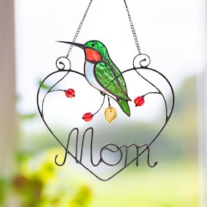 May include: A stained glass hummingbird decoration with a heart-shaped frame. The hummingbird is green, red, and white, perched on a wire frame with the word "Mom". Red berries and a gold leaf accent the design, suspended by a chain.