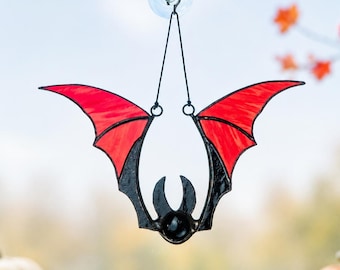 May include: A stained glass bat suncatcher featuring vibrant red wings with black outlines and a central black body. The bat is suspended by two thin black metal chains attached to a clear suction cup, designed for display on a window surface.