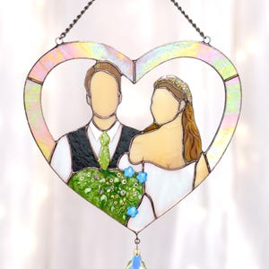 May include: Stained glass heart-shaped wedding art featuring a bride and groom. The couple is depicted with faceless figures, holding a bouquet. The heart has a rainbow iridescent border and is suspended by a chain. A crystal prism hangs below.