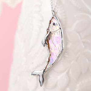 以下が含まれることがあります： 銀色の魚のペンダントネックレス。魚は、銀色のひれと小さな目を持つ、虹色に輝く体を持っています。ネックレスは繊細な銀色のチェーンから吊り下げられています。背景はソフトピンクと白です。