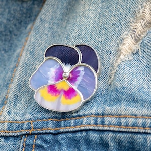 Könnte beinhalten: Eine silberfarbene Brosche mit einer bunten Glas-Stiefmütterchen-Blume mit violetten, gelben und rosa Blütenblättern. Die Brosche ist an einer Jeansjacke befestigt.