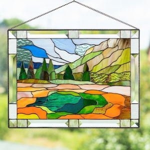 May include: A stained glass window depicting a landscape scene with a green lake, green trees, brown hills, and a blue sky with white clouds. The window is framed in a silver metal frame.