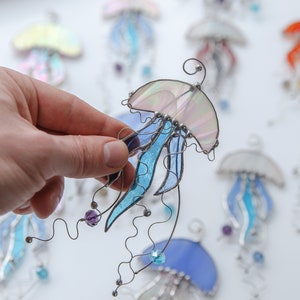 May include: Hand-held, iridescent stained glass jellyfish ornament with blue and purple accents. The jellyfish has a curved top, flowing tentacles, and decorative beads. Other jellyfish ornaments are visible in the background.