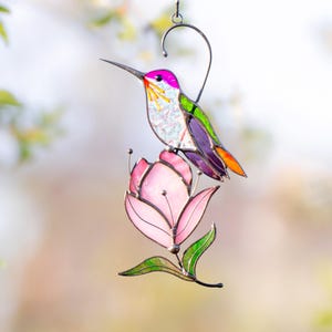 Stained glass hummingbird suncatcher with purple, green, and orange accents perched on a soft pink magnolia flower
