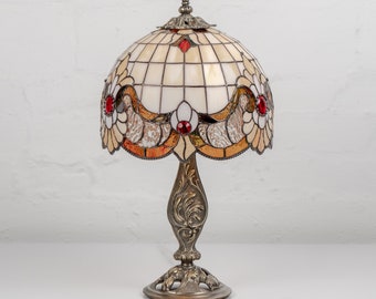 May include: A stained glass table lamp with a cream and brown patterned shade and red accents. The lamp has a metal base with an ornate design.
