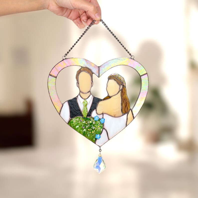 Hand holding a heart-shaped stained glass wedding portrait of a bride and groom, photographed indoors with soft warm background.