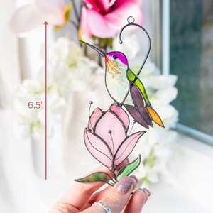 A hand holds a stained glass hummingbird with purple wings and green back perched on a pink magnolia, next to a window with floral decor and height marked as 6.5"