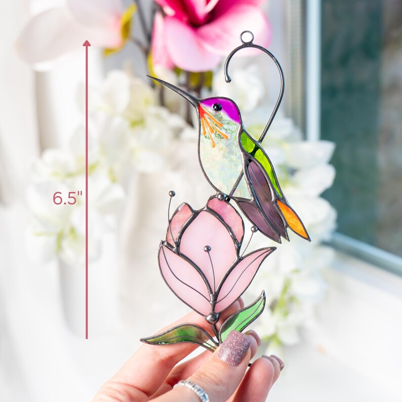 A hand holds a stained glass hummingbird with purple wings and green back perched on a pink magnolia, next to a window with floral decor and height marked as 6.5"