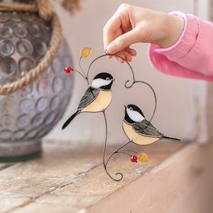 Stained glass bird suncatcher placed on wooden surface, handmade chickadee window hanging with metal branch and red berries, decorative glass bird art.
