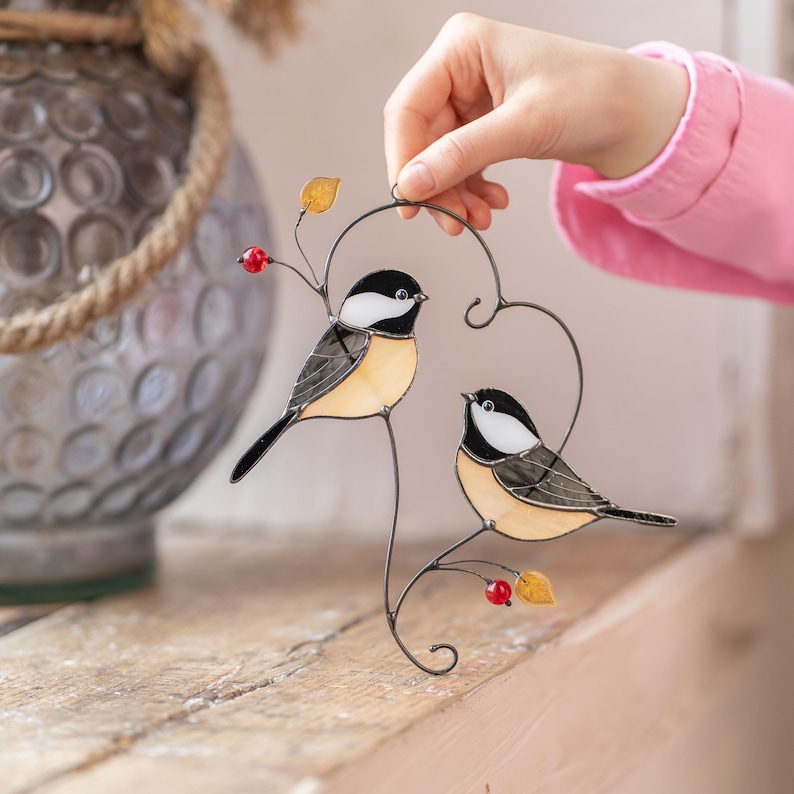 Stained glass bird suncatcher placed on wooden surface, handmade chickadee window hanging with metal branch and red berries, decorative glass bird art.
