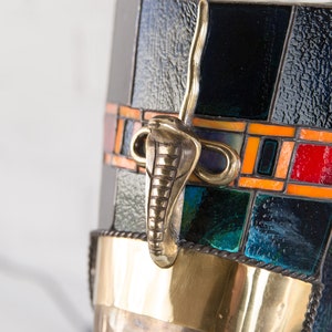 May include: A black and gold stained glass lamp with a cobra-shaped handle. The lamp has a geometric pattern with orange, red, and blue accents.