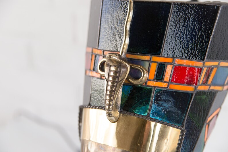 May include: A black and gold stained glass lamp with a cobra-shaped handle. The lamp has a geometric pattern with orange, red, and blue accents.