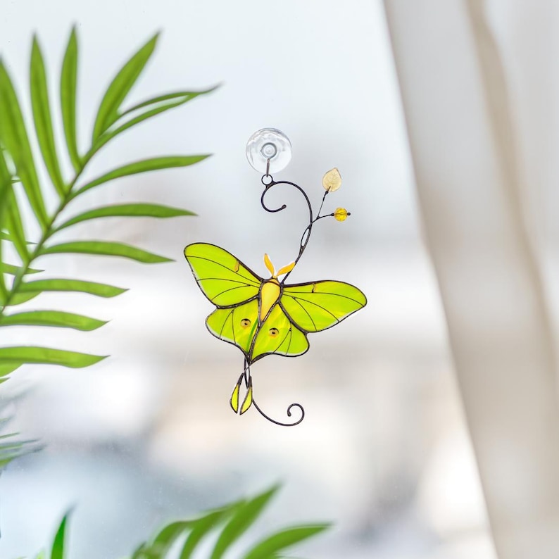 A vibrant green Luna Moth suncatcher made of stained glass, hanging on a window with a suction cup. The light illuminates the lime-green wings and delicate copper wire details. A blurred palm plant in the background enhances the garden decoration feel.