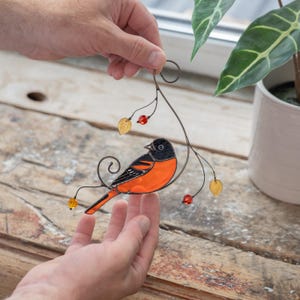 Close-up of a stained glass Baltimore oriole bird ornament in hands, with glass leaves and bead details.