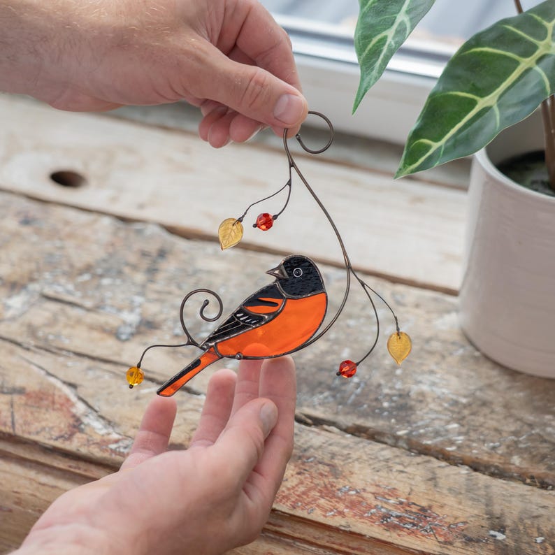 Close-up of a stained glass Baltimore oriole bird ornament in hands, with glass leaves and bead details.