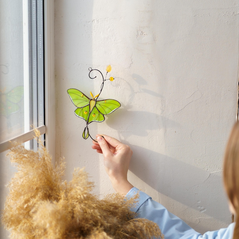 Close-up shots of a woman in a white sweater admiring a Luna Moth stained glass window hanging. The suncatcher features elegant wire curls and yellow glass accents, presented as a thoughtful gift idea for her or garden lovers.