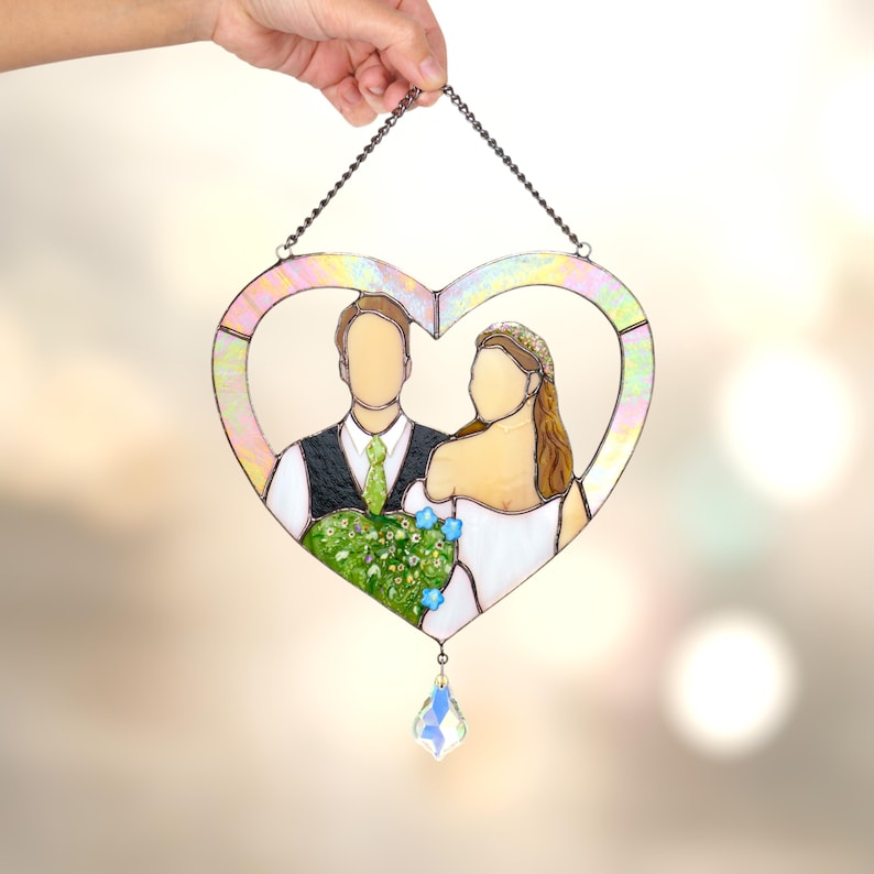 Hand holding a heart-shaped stained glass wedding portrait with iridescent frame and crystal drop against a soft glowing background.