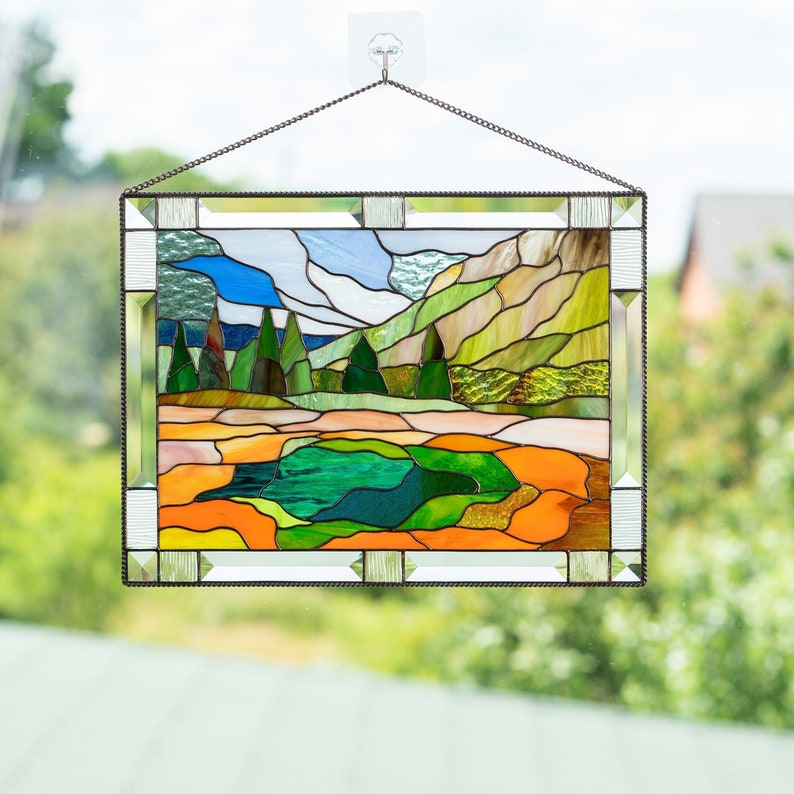 May include: A stained glass window depicting a landscape scene with a green lake, green trees, brown hills, and a blue sky with white clouds. The window is framed in a silver metal frame.