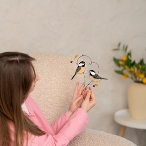 Woman holding stained glass bird suncatcher indoors, handmade chickadee window hanging, cozy home decor with nature inspired glass birds.