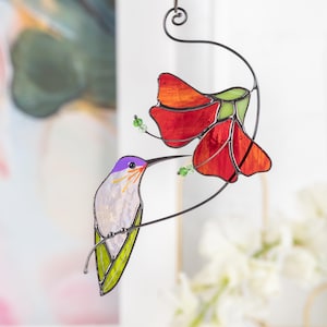Stained glass hummingbird suncatcher drinking nectar from a red flower, hanging in a bright window.