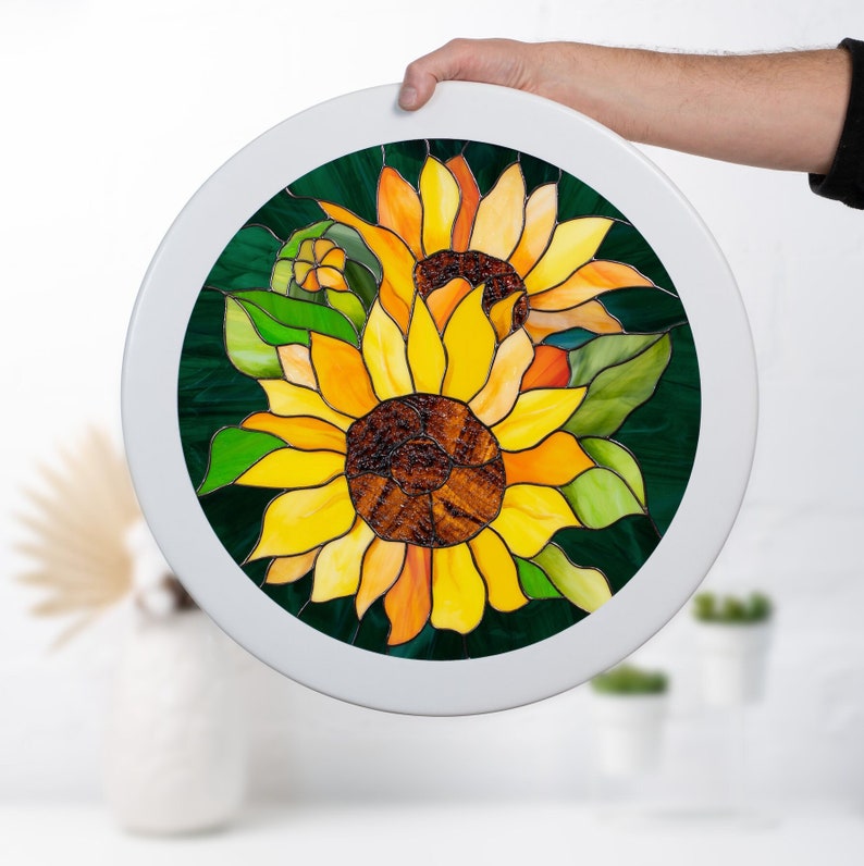 May include: A round stained glass panel featuring two sunflowers with yellow petals, brown centers, and green leaves on a dark green background. The panel is framed in white.