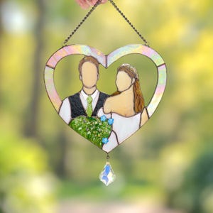Hand holding a heart-shaped stained glass wedding portrait of a bride and groom with a crystal drop, photographed outdoors with blurred greenery.