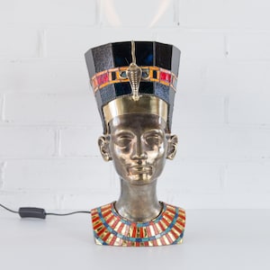 May include: A bronze bust of an Egyptian queen wearing a black and gold headdress with colorful mosaic details. The bust is a table lamp with a cord and switch.