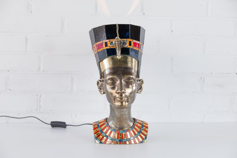 May include: A bronze bust of an Egyptian queen wearing a black and gold headdress with colorful mosaic details. The bust is a table lamp with a cord and switch.