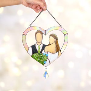 Heart-shaped stained glass wedding portrait with iridescent frame and crystal drop held by hand in front of a bright bokeh background.